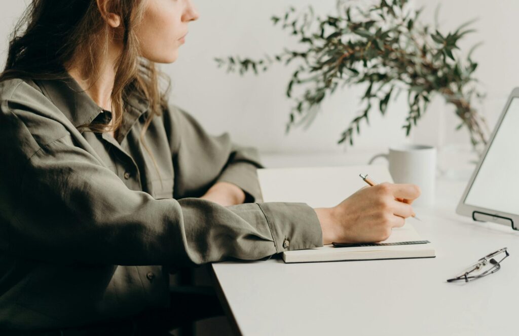 Freelancer writing notes in a minimalist home office with a white tablet, plant, and reading glasses.